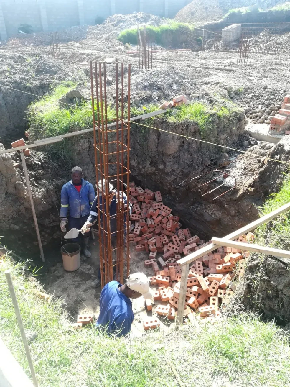 Project site — deep foundation pit with brick footing and rebar cage, workers in background