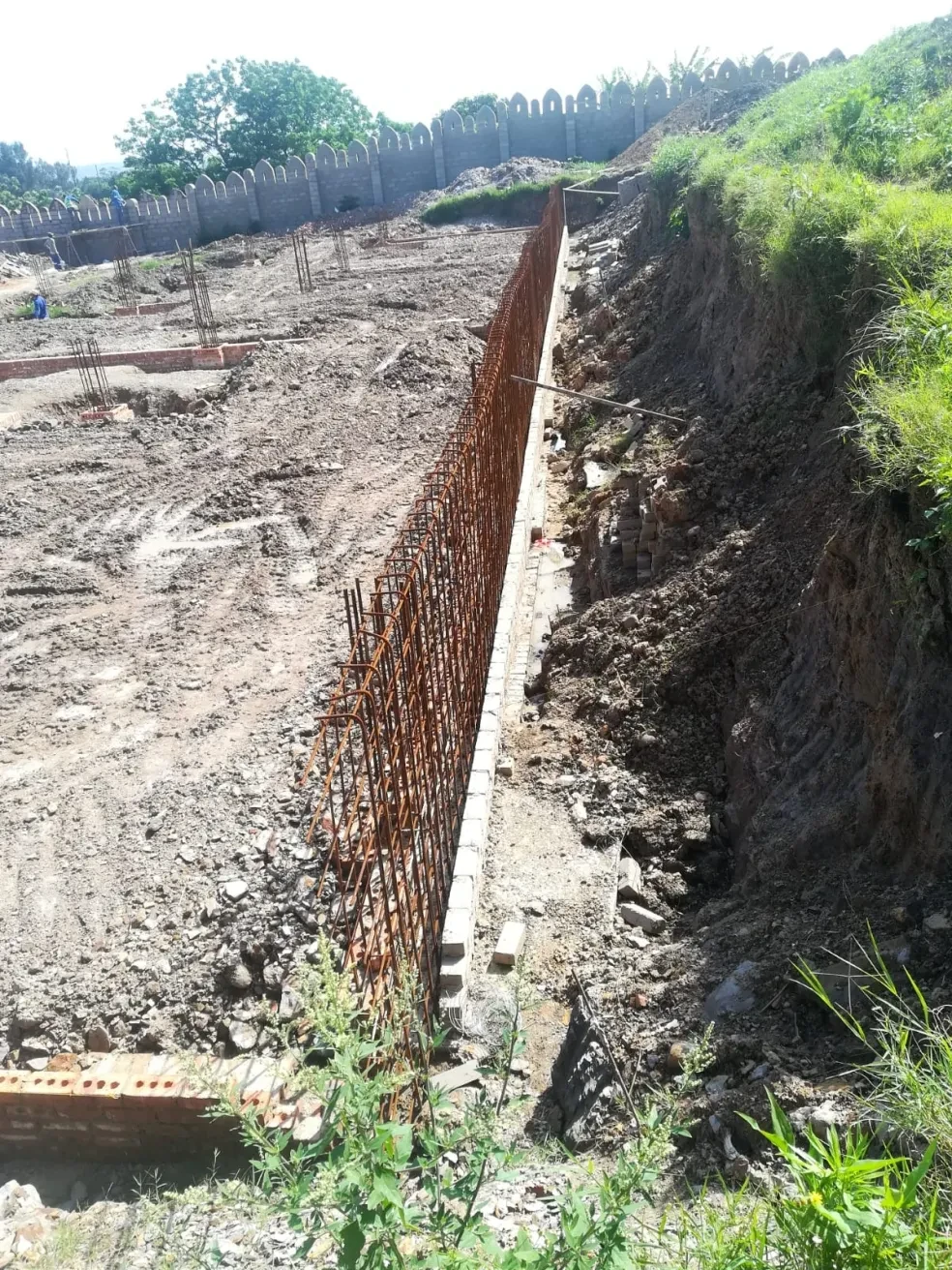 Project site — long rebar wall foundation trench with crenellated boundary wall on hillside