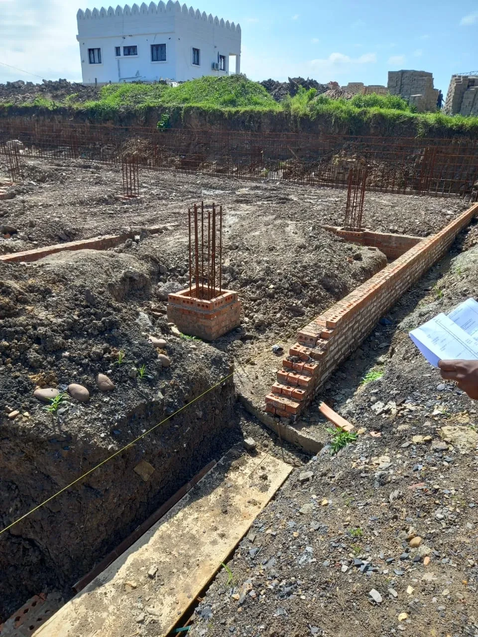 Project site — wide foundation excavation with rebar columns, brick footings and crenellated wall with palm trees
