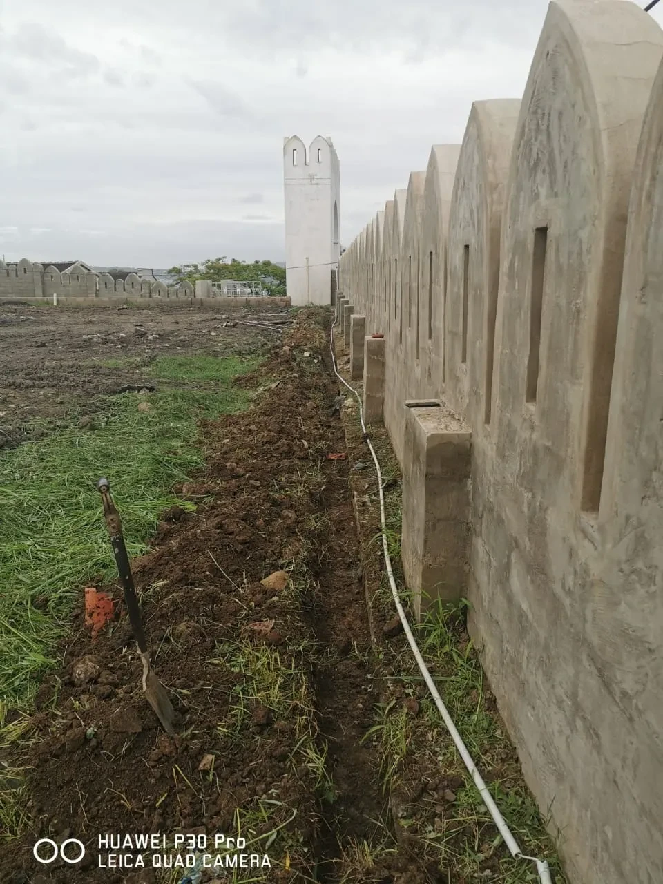 Project site — crenellated perimeter wall with arched panels and utility conduit trench being dug alongside