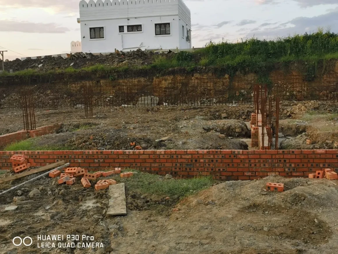 Project site — brick foundation wall with rebar columns and white crenellated building in background at dusk