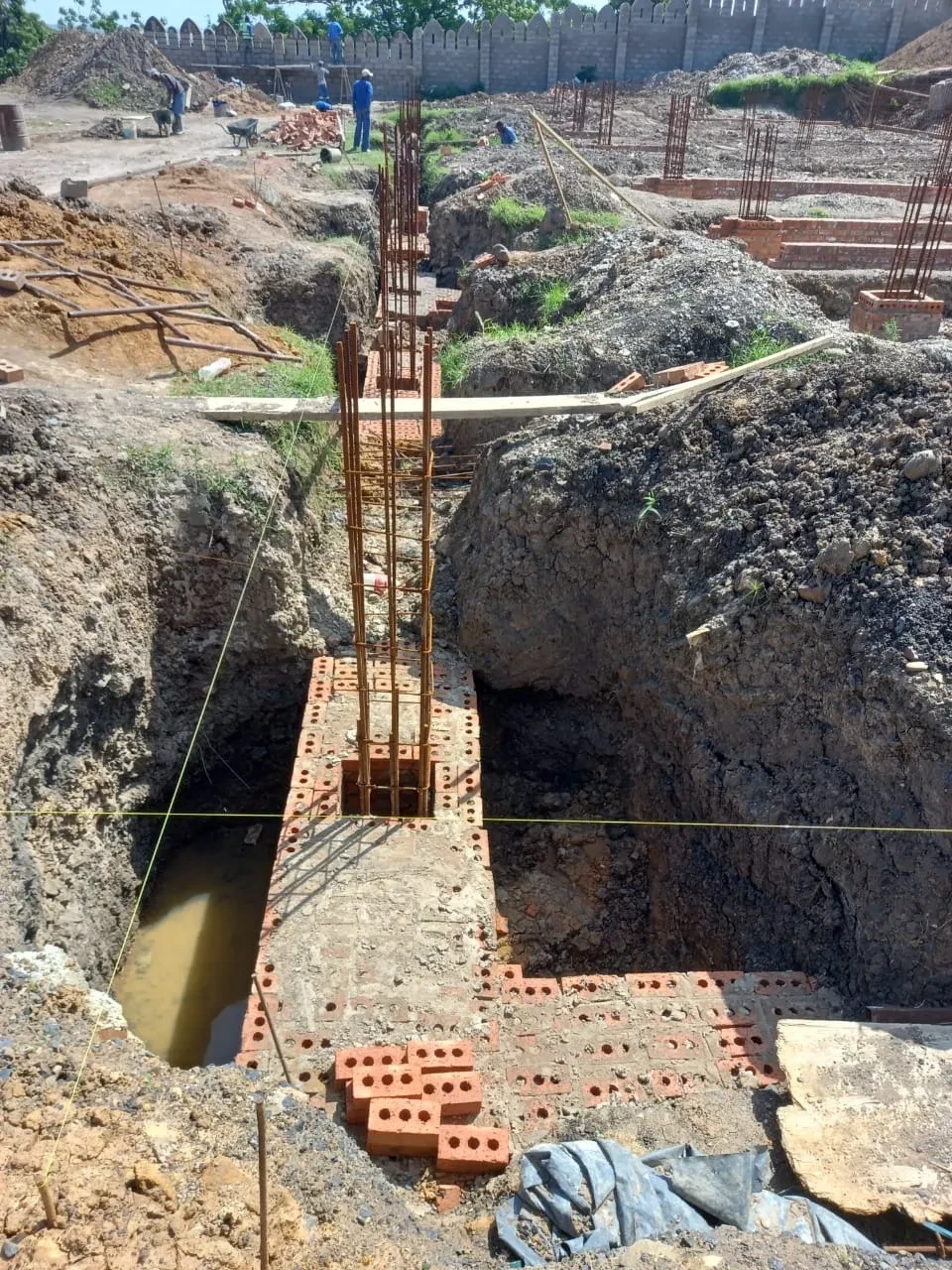 Project site — deep foundation trench with tall rebar cage columns and brick footings, crenellated wall in background