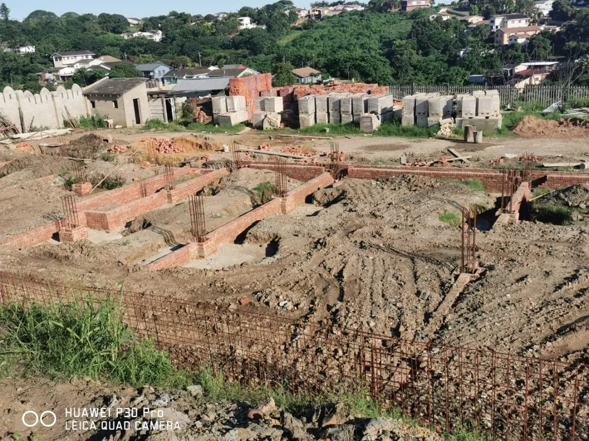 Project site — wide foundation excavation with rebar columns, brick footings and workers in background with crenellated perimeter wall