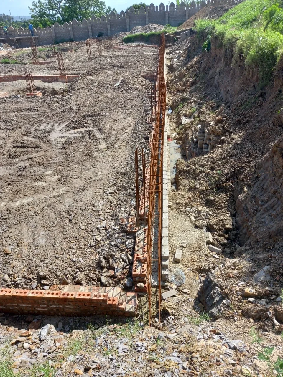 Project site — deep foundation trench with rebar cage and brick column footings, crenellated wall and white tower in background