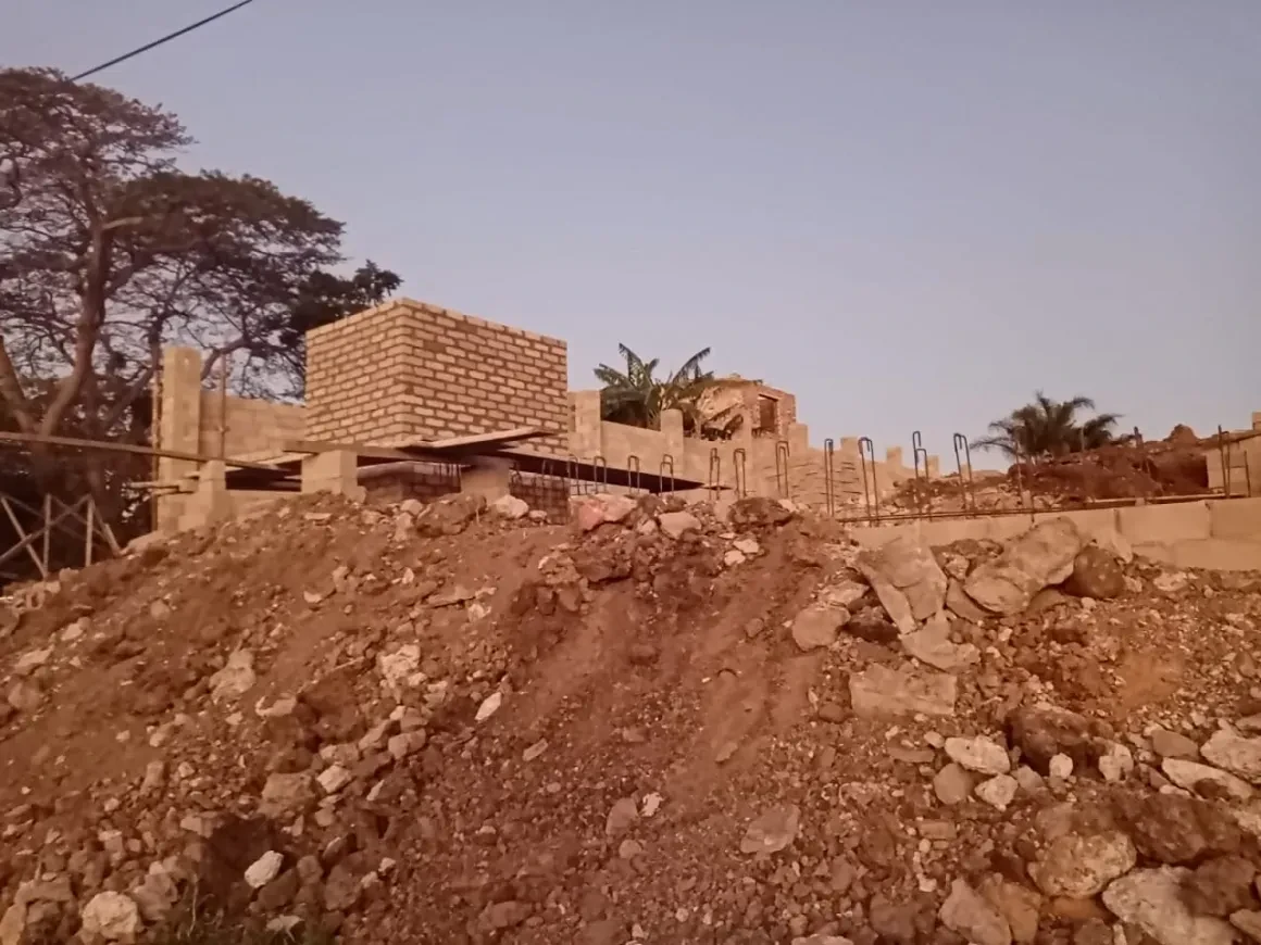 Project site — brick walls and crenellated parapet rising from red earth excavation mound at dusk