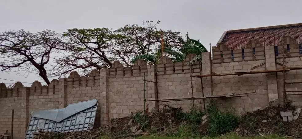 Project site — crenellated concrete block perimeter wall with scaffolding, bare trees and tiled roof building behind