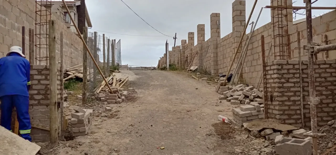 Project site — long construction road between concrete block walls with rebar columns and worker in blue overalls