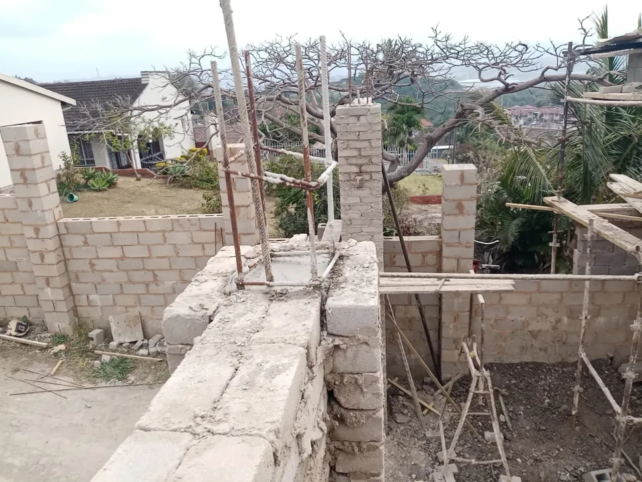 Project site — brick and concrete block walls with rebar columns, bare trees and Durban suburb in background
