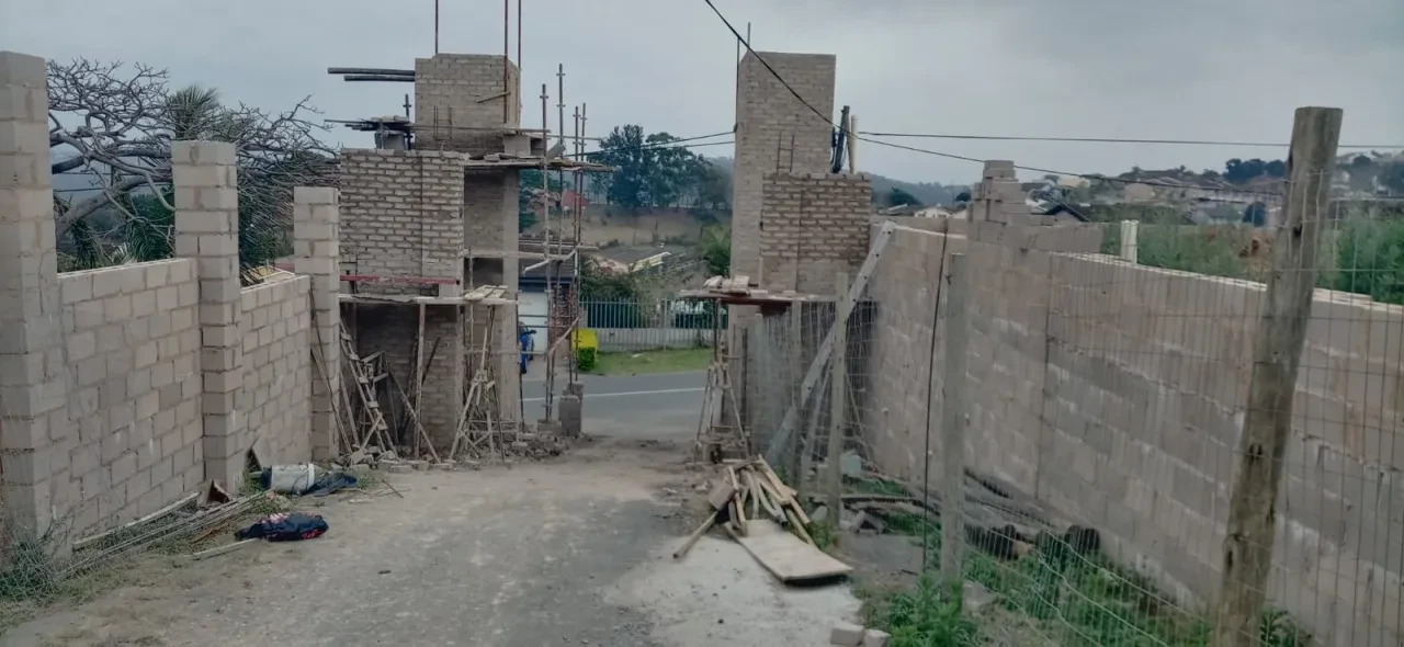 Project site — concrete block gateway under construction with scaffolding, road and green fence visible beyond