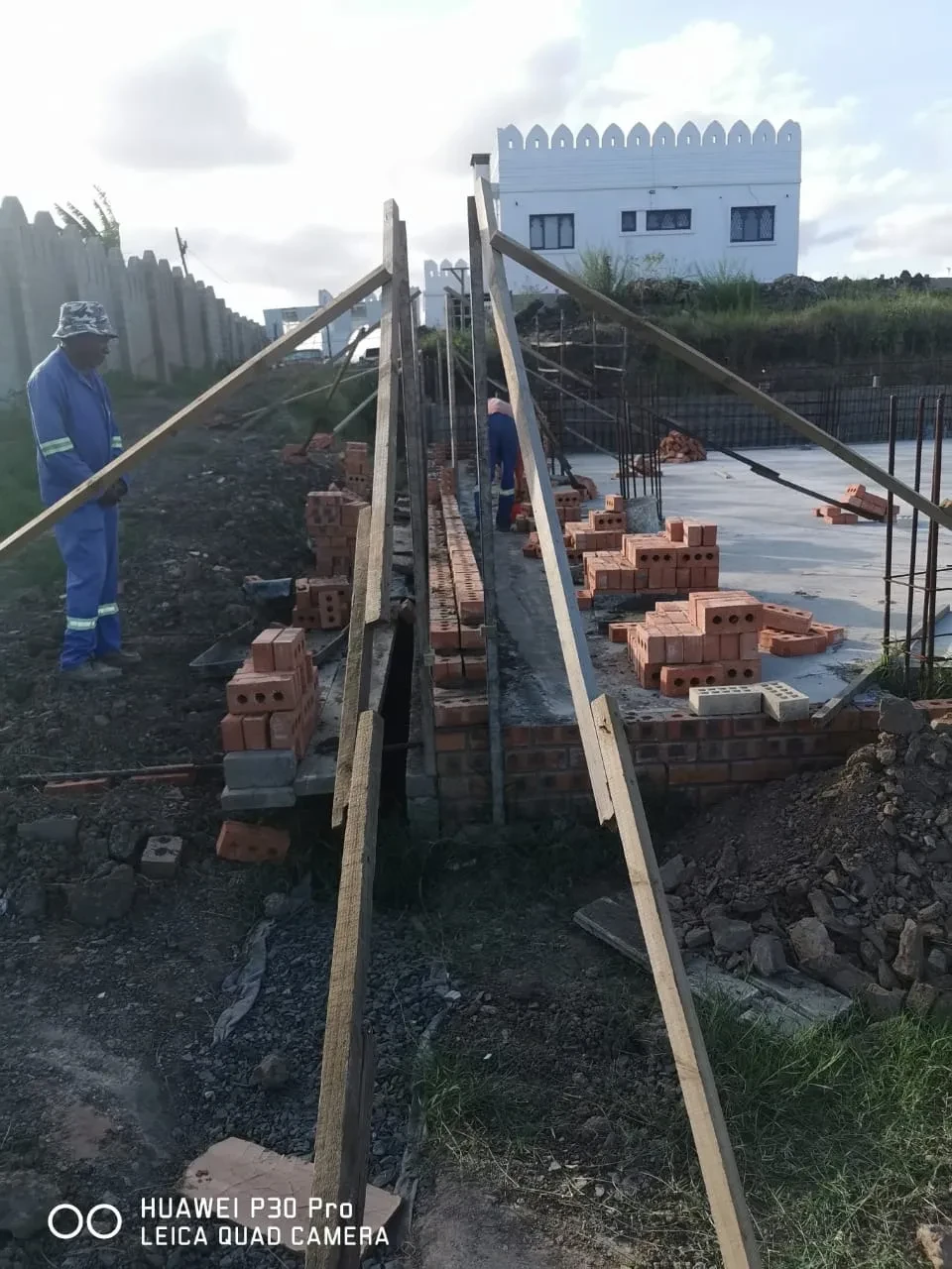 Project site — workers laying brick foundation wall on concrete slab with white crenellated tower in background