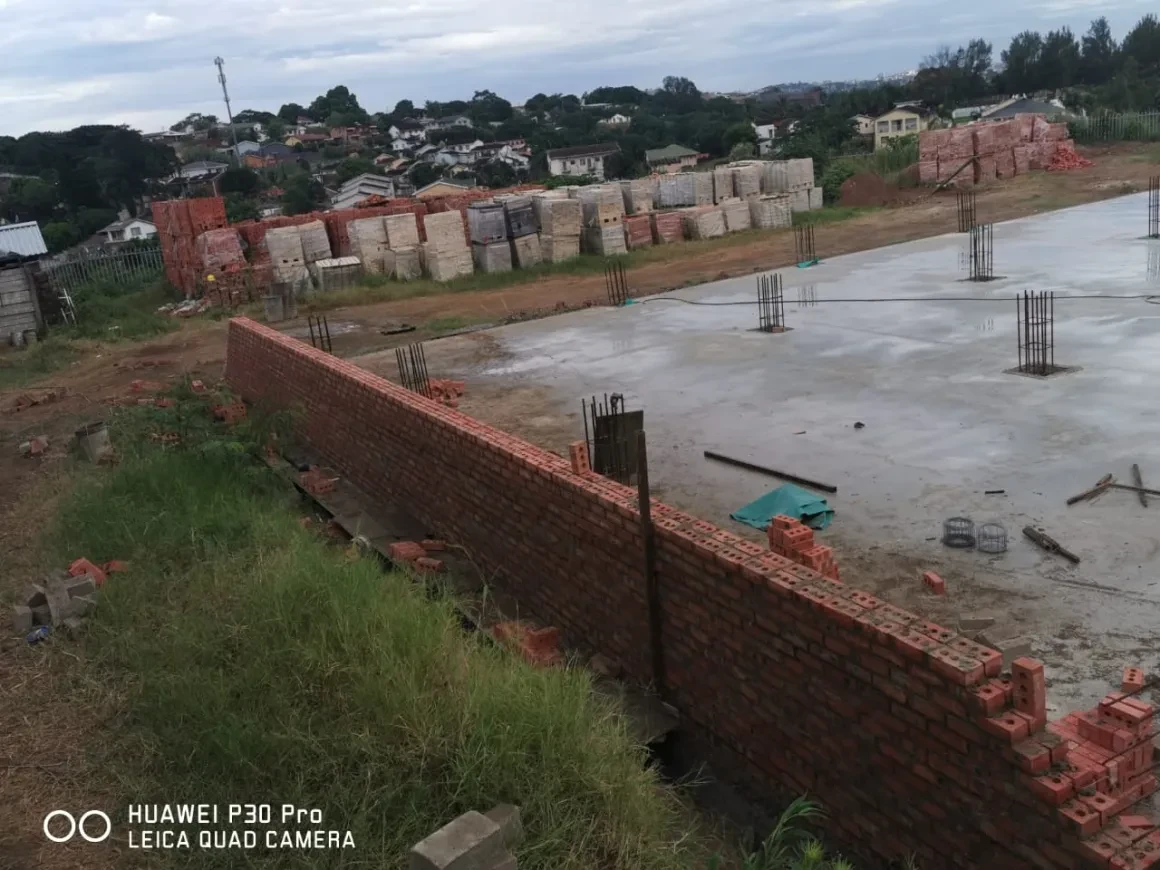 Project site — brick foundation wall with rebar columns on concrete slab, crenellated perimeter wall with palm trees