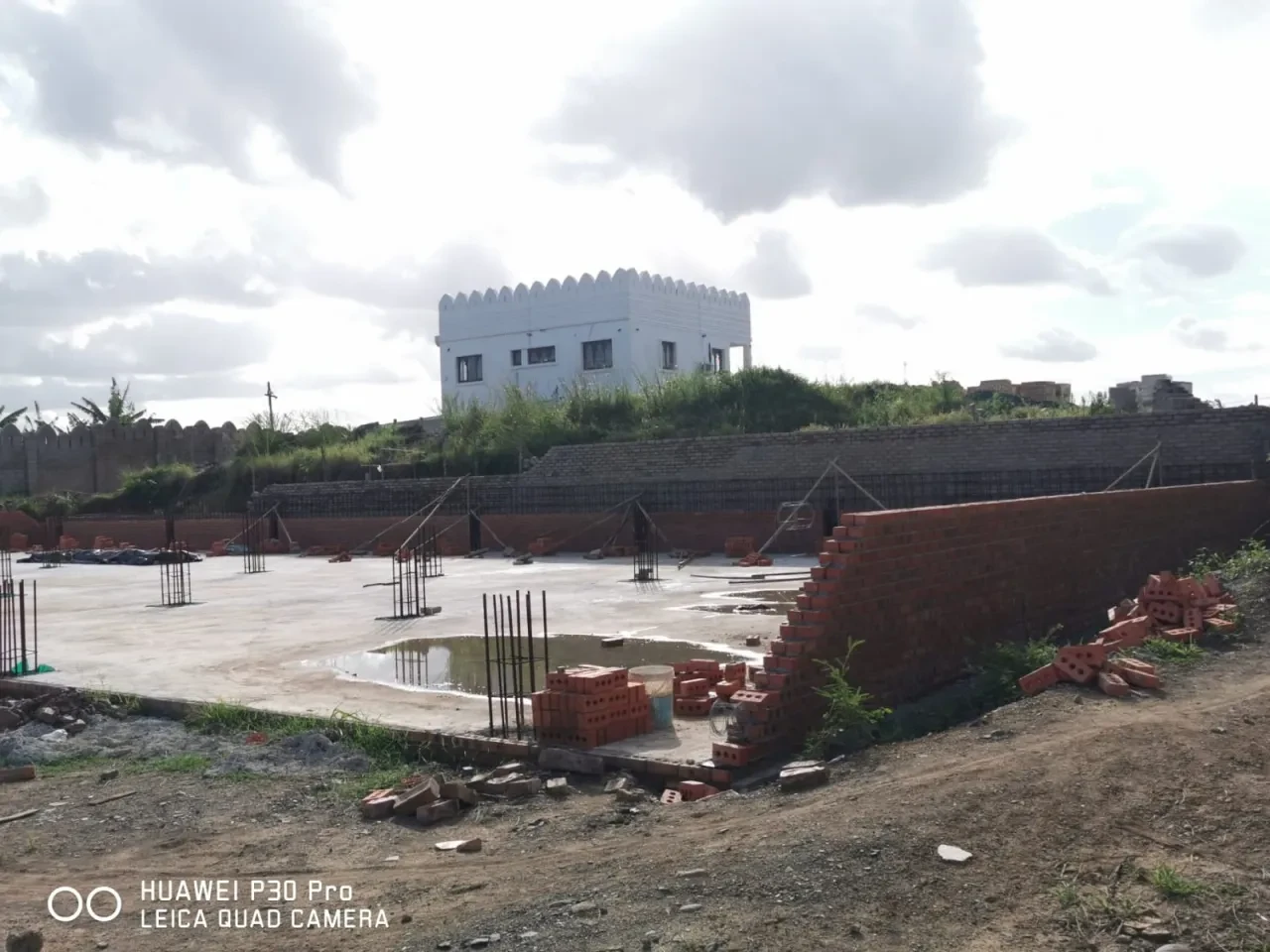 Project site — freshly poured concrete slab with rebar columns, brick retaining walls and white crenellated building in background