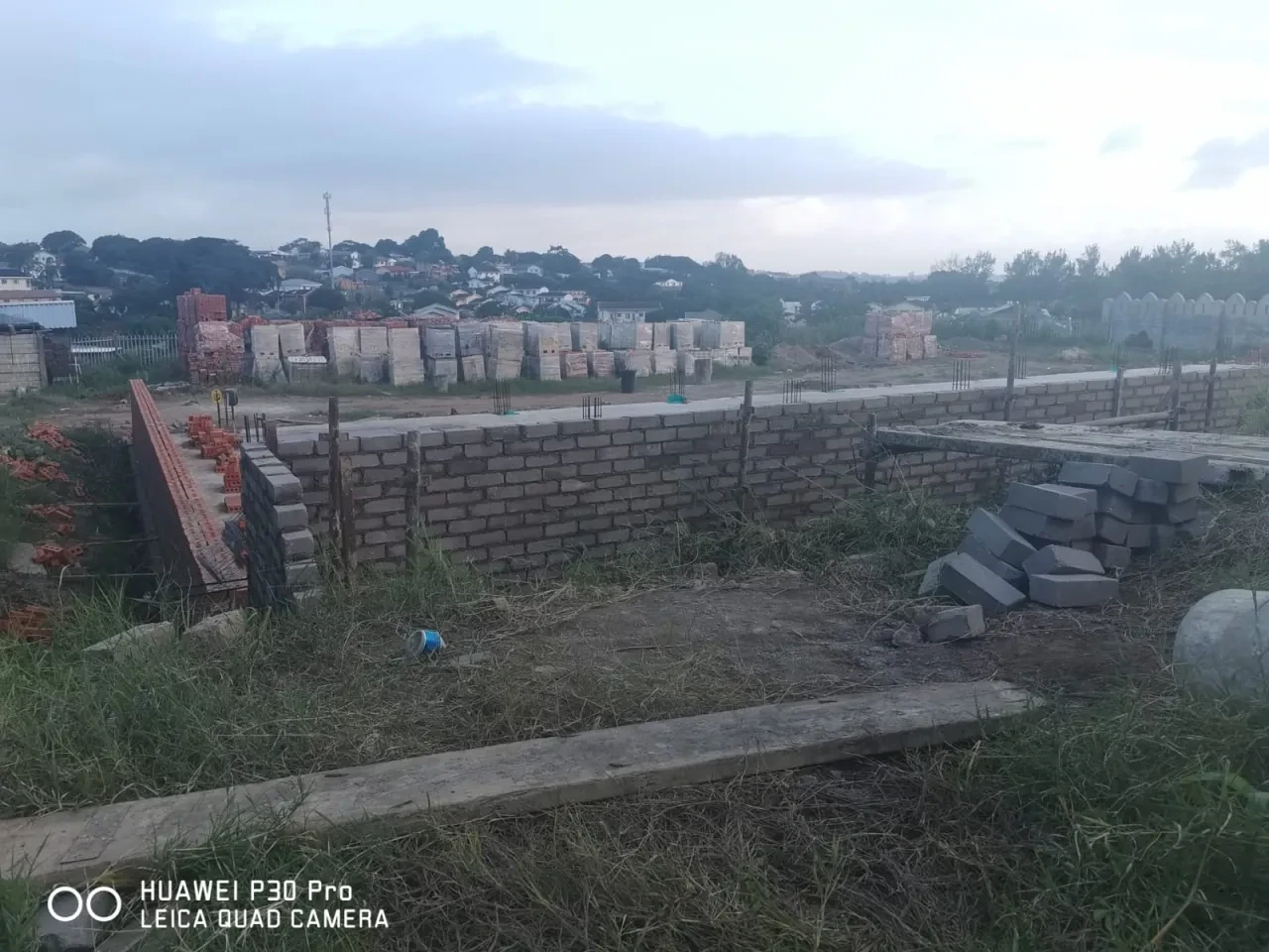 Project site — concrete block foundation walls with rebar columns, stacked building materials and Durban suburb in background at dusk