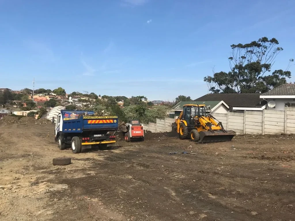 Project site — cleared site with dump truck, bobcat and JCB backhoe loader, Durban suburb in background