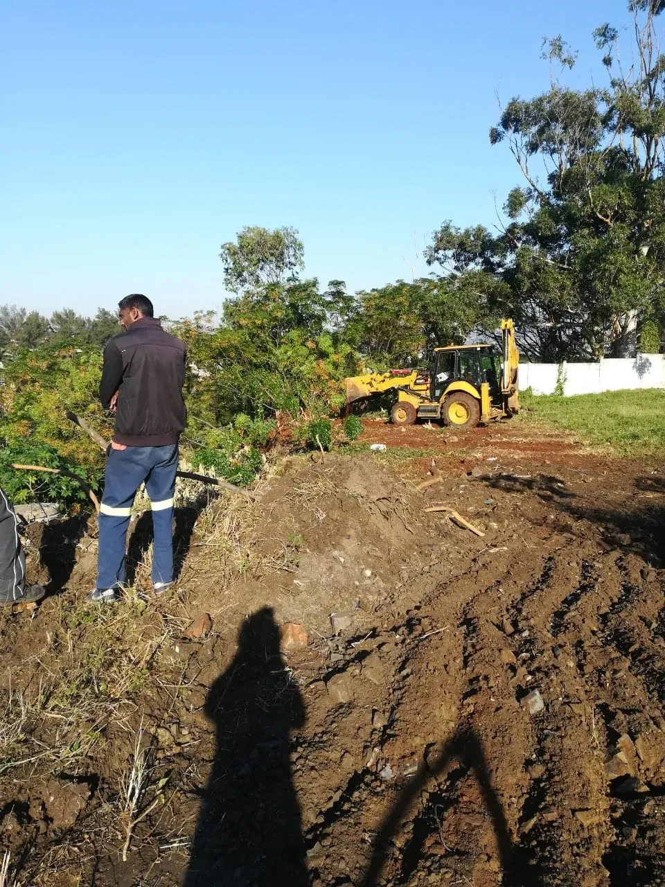 Project site — worker in hi-vis trousers overseeing yellow backhoe excavation on cleared site with white perimeter wall