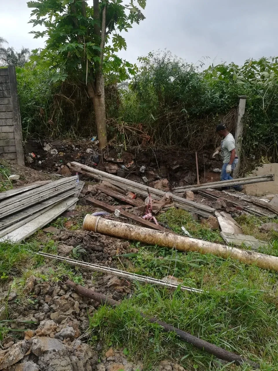 Project site — utility trench dug alongside brick house with banana trees and concrete fence posts