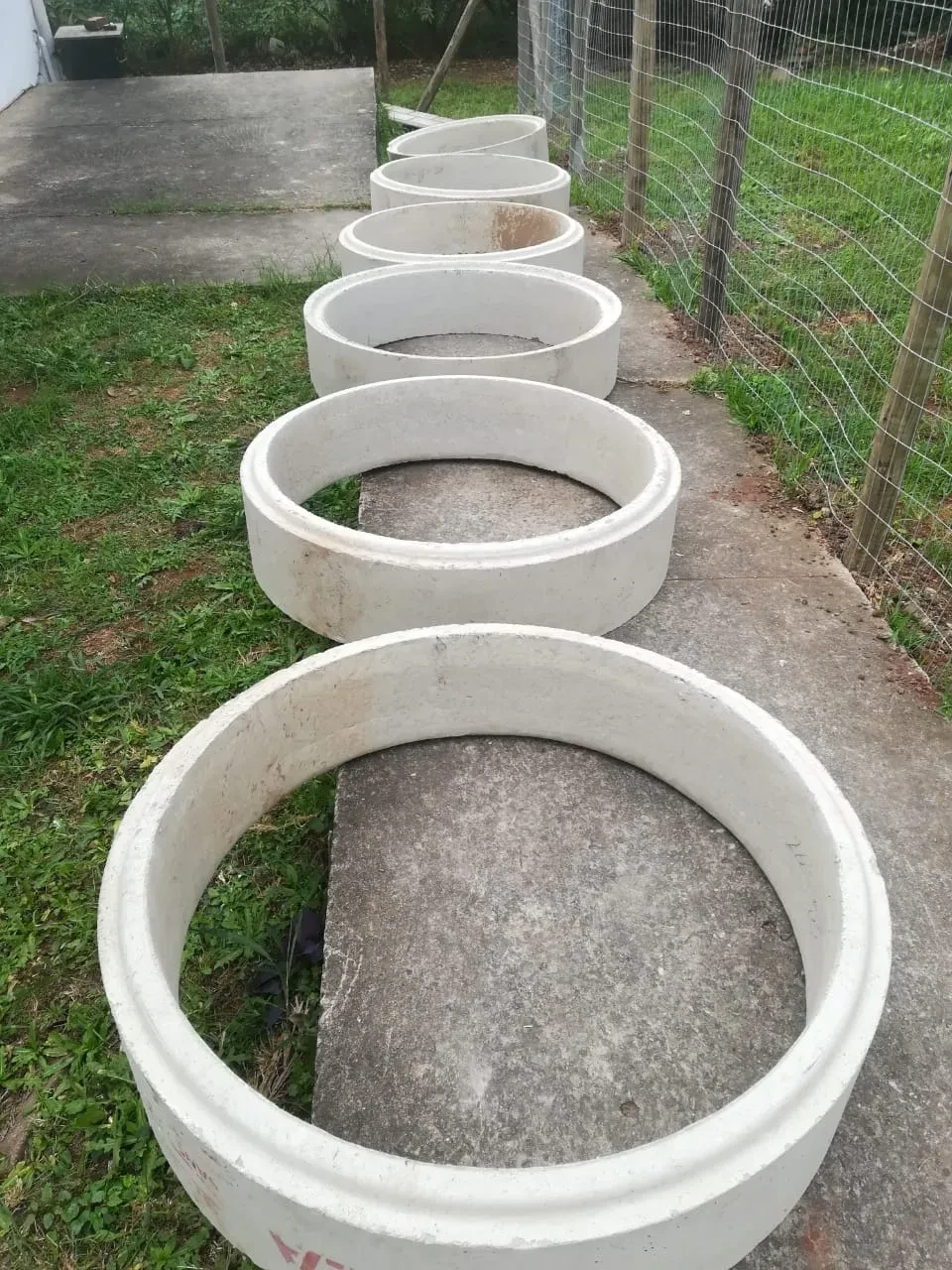 Project site — row of concrete manhole rings lined up on paved path beside wire fence and grass